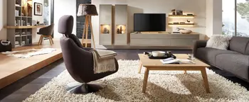 Wohnzimmer mit grauem Sessel im Vordergrund, hellem TV-Möbel, Couchtisch und Bücherregal auf hellem Hochflorteppich.